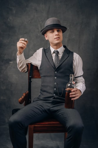 Homme assis sur une chaise, portant un costume gris avec une cravate et un chapeau, tenant une bouteille et un verre de whisky, avec un pistolet attaché au dossier de la chaise, sur un fond sombre.