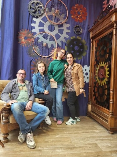 Quatre personnes souriantes, deux adultes et deux enfants, posant dans une pièce décorée de grandes roues dentées métalliques accrochées au mur et d'une grande horloge en bois à droite.
