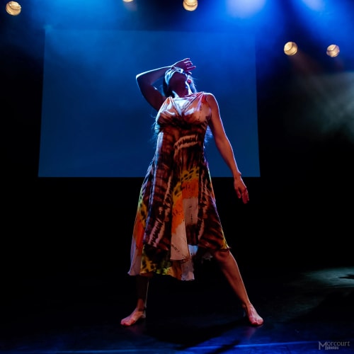 Femme dansant sur scène sous un éclairage bleu avec une robe colorée, posant avec un bras sur le front et l'autre tendu vers le bas.