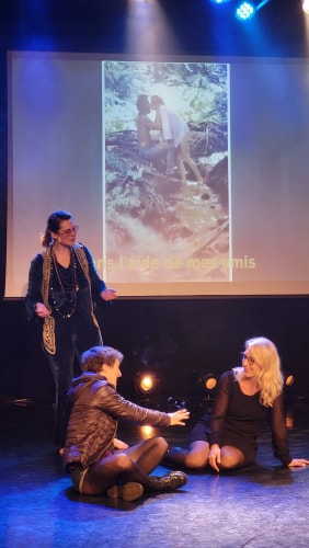 Trois femmes sur une scène, deux assises au sol en train de discuter et une debout, en arrière-plan un écran avec une image floutée de deux personnes s'embrassant dans un décor naturel.