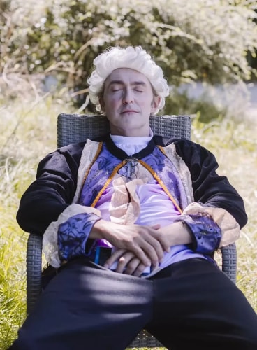 Homme portant une perruque blanche et un costume d'époque assis les yeux fermés sur une chaise extérieure dans un jardin.