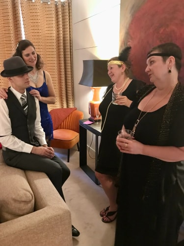 Quatre personnes habillées de façon élégante dans un salon, un homme portant un chapeau assis sur un canapé, une femme en robe bleue se tenant derrière lui, et deux femmes en robe noire tenant des verres et discutant.