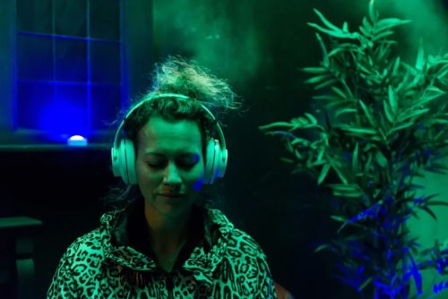 Personne portant un casque audio blanc, les yeux fermés, dans une pièce sombre éclairée de lumière verte et bleue avec une plante en arrière-plan