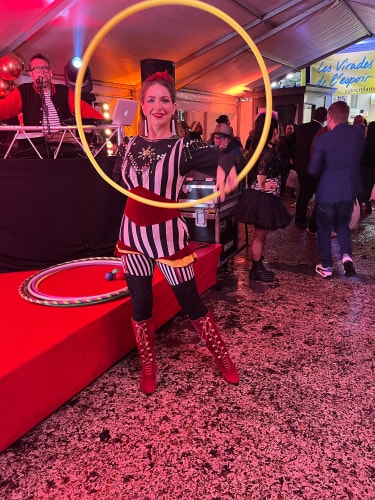 Femme artiste de cirque vêtue d'un costume rayé noir et blanc avec des bottes rouges, manipulant un grand cerceau jaune dans une tente animée avec un DJ et des spectateurs en arrière-plan.