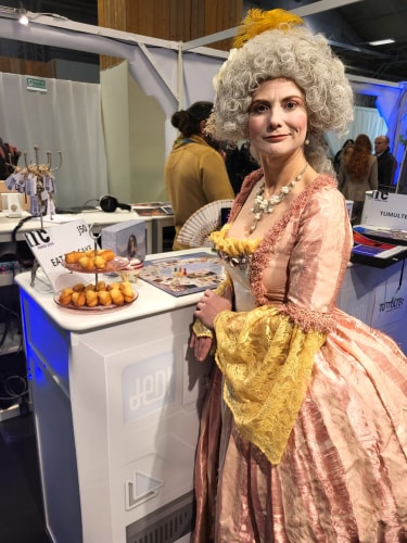 Femme en costume d'époque rose et jaune avec perruque grise bouclée, posant devant un stand avec des petits gâteaux sur un présentoir à étages.