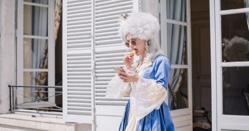 Une personne en costume d'époque, pour l'expérience Versailles d'In the Cake, avec une perruque blanche bouclée, portant des lunettes de soleil et mangeant tout en regardant son téléphone devant une maison avec des portes-fenêtres blanches.