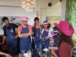Groupe de six adultes portant des tabliers bleus et des chapeaux colorés, participant à une activité de préparation de boissons autour d'une table avec des ingrédients et des ustensiles de bar.