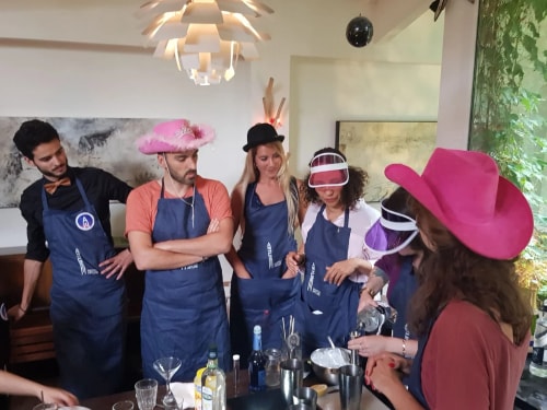 Groupe de six adultes portant des tabliers bleus et des chapeaux colorés, participant à une activité de préparation de boissons autour d'une table avec des ingrédients et des ustensiles de bar.