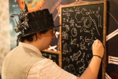 Personne portant un chapeau orné d'une décoration et des lunettes, écrivant des formules scientifiques et des dessins sur un tableau noir.