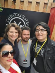 Groupe de quatre personnes souriantes portant des badges jaunes autour du cou, posant devant une bannière noire avec le logo et le texte "World Experience", dans un cadre extérieur avec une palissade en bois.