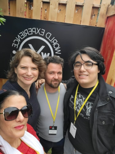 Groupe de quatre personnes souriantes portant des badges jaunes autour du cou, posant devant une bannière noire avec le logo et le texte "World Experience", dans un cadre extérieur avec une palissade en bois.