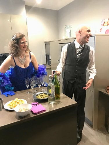 Deux personnes en tenue élégante discutent dans une cuisine moderne, une femme en robe bleue et un homme en costume noir avec une cravate, debout près d'un plan de travail avec des verres à champagne, des olives, des amuse-gueules et des bouteilles.