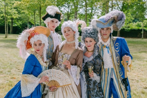 Groupe de cinq personnes habillées en costumes d'époque du XVIIIe siècle avec perruques blanches et chapeaux décorés, posant dans un jardin, certaines tenant des verres à la main et une éventail.