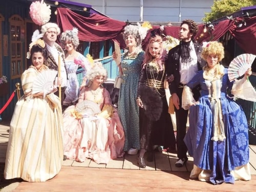 Groupe de huit personnes habillées en costumes d'époque rococo, portant des perruques, des robes élaborées et tenant des éventails, posant sur une terrasse en bois devant un bâtiment avec un décor de tissu rouge.