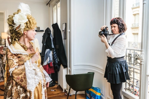 Une femme en costume d'époque doré avec un éventail pose tandis qu'une autre femme en tenue moderne prend une photo avec un appareil photo dans une pièce lumineuse avec une grande fenêtre.