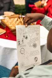 Personne tenant une feuille de papier avec des dessins techniques et textes manuscrits, en arrière-plan un jeu d'échecs flou sur une table.