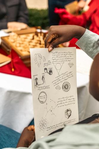 Personne tenant une feuille de papier avec des dessins techniques et textes manuscrits, en arrière-plan un jeu d'échecs flou sur une table.