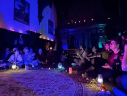 Un groupe de personnes assises en cercle sur le sol dans une pièce faiblement éclairée avec des bougies, des lanternes et des décorations, participant à une activité ou cérémonie dans une ambiance chaleureuse et contemplative.