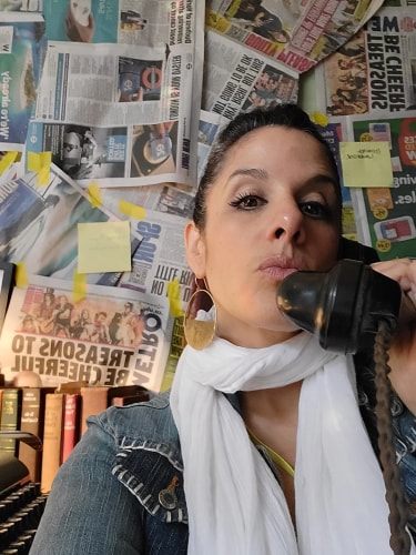 Femme portant une écharpe blanche et une veste en jean, tenant un ancien combiné téléphonique près de son visage, avec un mur couvert de journaux et de notes en arrière-plan.