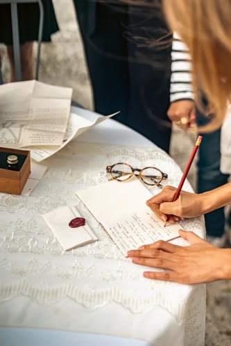 Une personne écrit une lettre à la main sur une table recouverte d'une nappe blanche brodée, avec des lunettes posées à côté, une enveloppe cachetée et des papiers éparpillés.
