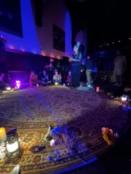 Une femme habillée de façon élégante se tient debout au centre d'un tapis rond, entourée de personnes assises en cercle dans une pièce sombre avec des lumières tamisées, des lampes et des bougies disposées autour du tapis, créant une ambiance intime et mystérieuse.