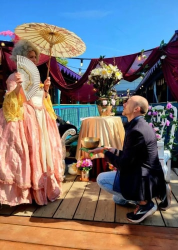 Une femme en robe rose d'époque tient un éventail et un parasol, un homme à genoux face à elle tient un bol en métal dans un décor extérieur décoré avec des fleurs et des rideaux pourpres.