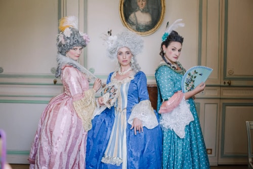 Trois femmes en costumes d'époque du XVIIIe siècle, avec des robes élégantes et des coiffures ornées de plumes, posant dans une pièce décorée à l'ancienne avec un tableau encadré au mur.