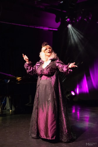 Une femme sur scène en robe longue violette avec des manches bouffantes, éclairée par des projecteurs violets, chantant ou récitant avec expression théâtrale.