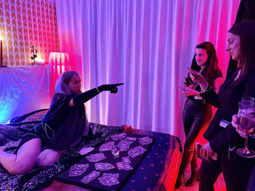 Une femme en costume spirituel pointe du doigt tandis qu'elle est allongée sur un lit décoré avec des cartes et une boule de cristal, devant deux autres femmes debout qui tiennent des verres et des cartes, dans une pièce éclairée par des lumières roses et bleues avec un rideau blanc en arrière-plan.