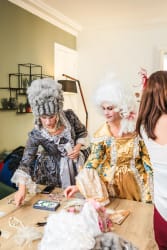 Deux femmes en costumes d'époque avec perruques blanches et robes ornées, examinant des bijoux posés sur une table en bois dans une pièce lumineuse.