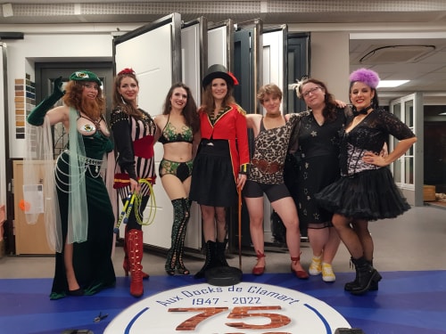 Groupe de sept femmes en costumes colorés et variés posant ensemble dans une pièce intérieure, certaines avec des accessoires comme un chapeau haut de forme, un fouet et des bottes.
