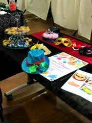 Table couverte d'une nappe noire avec plusieurs chapeaux colorés et pailletés, masques vénitiens, un présentoir à gâteaux avec des pâtisseries et des papiers décoratifs colorés.