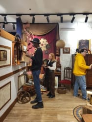 Trois personnes dans une pièce décorée de style vintage avec des objets anciens, une femme rousse tenant un objet, un homme portant un chapeau noir examinant un livre, et une autre personne en sweat jaune marchant vers la droite.