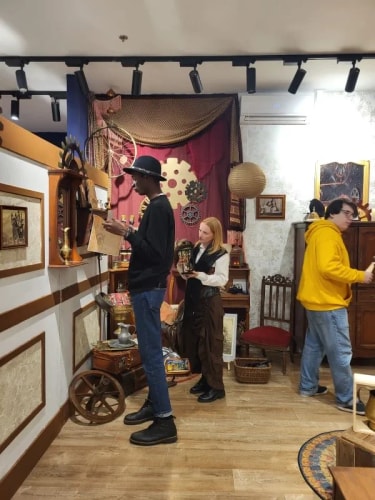Trois personnes dans une pièce décorée de style vintage avec des objets anciens, une femme rousse tenant un objet, un homme portant un chapeau noir examinant un livre, et une autre personne en sweat jaune marchant vers la droite.