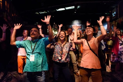 Un groupe de personnes souriantes lève les mains en l'air dans un environnement intérieur sombre, exprimant joie et enthousiasme.