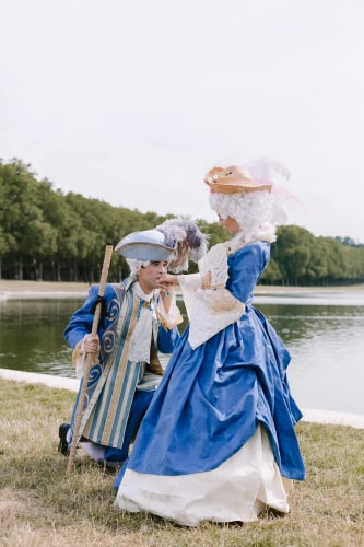 Un homme en costume bleu et chapeau avec plumes embrasse la main d'une femme en robe bleue et perruque blanche assise près d'un plan d'eau et d'arbres.