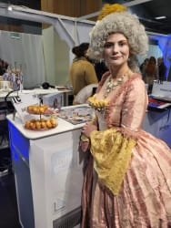 Femme en costume d'époque avec une perruque grise bouclée et une robe rose et jaune, posant près d'un stand avec des pâtisseries sur un présentoir à étages.