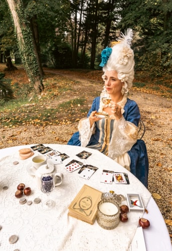 Une femme habillée en costume historique bleu avec une perruque blanche et des ornements, assise à une table ronde blanche en dentelle à l'extérieur, tenant une tasse de thé dorée, entourée de cartes à jouer, d'une théière, de livres et d'autres petits objets, en forêt en automne.