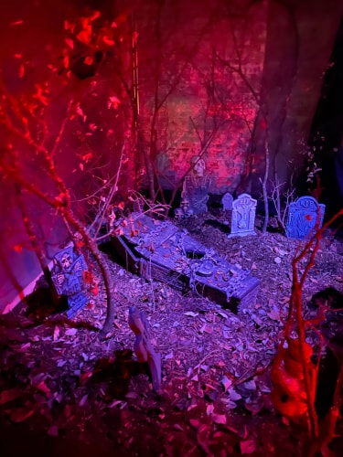 Scène de cimetière miniature avec plusieurs pierres tombales, un cercueil au centre, des squelettes et des branches d'arbres dégarnies, éclairage rouge et violet créant une ambiance effrayante.