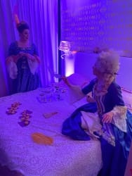Deux personnes en costumes d'époque assises et debout près d'un lit recouvert d'une nappe en dentelle, jouant aux échecs et tenant des éventails, dans une pièce éclairée par une lumière violette.