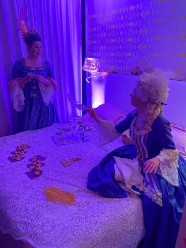 Deux personnes en costumes d'époque assises et debout près d'un lit recouvert d'une nappe en dentelle, jouant aux échecs et tenant des éventails, dans une pièce éclairée par une lumière violette.