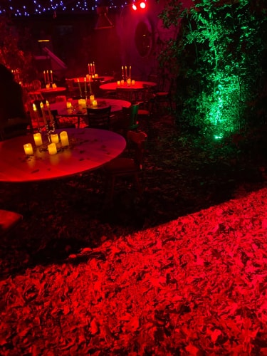 Tables rondes avec chaises disposées à l'extérieur, décorées de bougies jaunes, éclairage rouge et vert créant une ambiance chaleureuse et mystérieuse, feuilles mortes au sol.