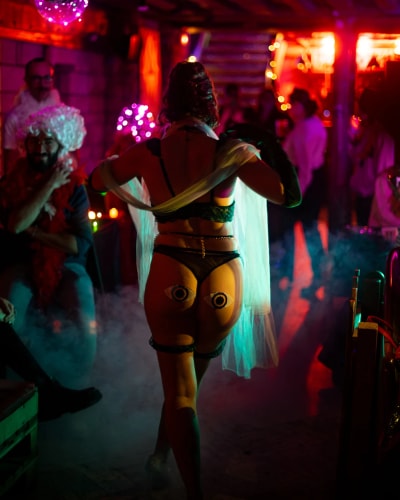 Personne en tenue légère noire avec des décorations en forme d'yeux sur les fesses, dans une ambiance de fête avec des lumières colorées et des spectateurs en arrière-plan.