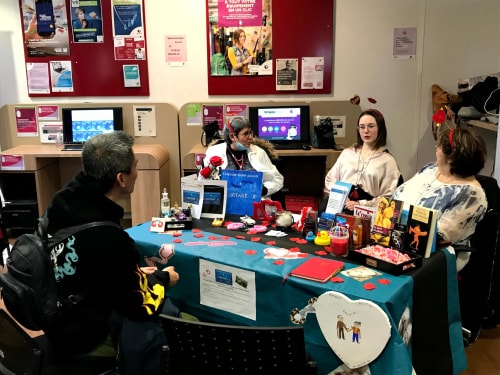 Quatre personnes discutant autour d'une table décorée avec des objets, des livres et des décorations en forme de cœur dans une salle avec des panneaux d'information affichés sur les murs.