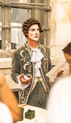 Homme en costume d'époque avec perruque bouclée et maquillage, tenant un papier et parlant devant une fenêtre ancienne, avec des personnes floues au premier plan.