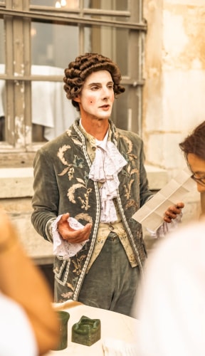 Homme en costume d'époque avec perruque bouclée et maquillage, tenant un papier et parlant devant une fenêtre ancienne, avec des personnes floues au premier plan.