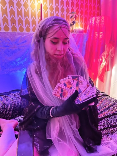 Femme voilée et gantée tenant un éventail de cartes de tarot dans un cadre coloré avec des bougies allumées en arrière-plan.