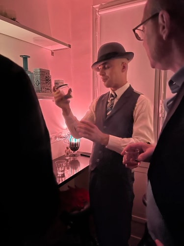 Un homme portant un gilet, une chemise blanche, une cravate et un chapeau, tenant un verre et semblant examiner son contenu dans une pièce éclairée par une lumière rouge, avec deux autres personnes partiellement visibles autour de lui.