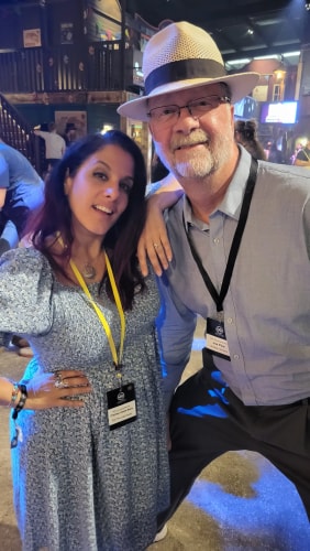 Une femme et un homme souriants portant des badges, la femme en robe bleue à motifs floraux avec un collier et plusieurs bracelets, l'homme en chemise bleue claire et chapeau blanc, posant dans un lieu intérieur avec des éclairages colorés et d'autres personnes en arrière-plan.