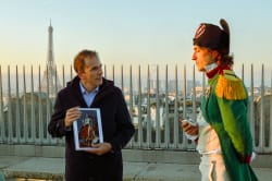 Deux hommes debout sur une terrasse en extérieur avec la Tour Eiffel en arrière-plan. L'un porte un costume historique de style militaire napoléonien et tient un objet dans la main, l'autre homme tient une photo d'un portrait de Napoléon.